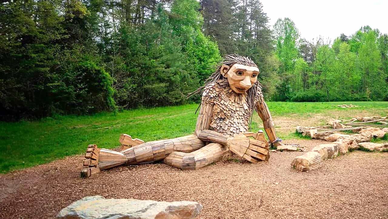 This image depicts a large wooden sculpture situated in a natural, outdoor setting. The sculpture represents a seated figure with exaggerated, whimsical features, crafted from logs and branches. The figure has a long, pointed nose, large eyes, and a body made of segmented wood pieces, giving it a rustic, handcrafted appearance. The surrounding area includes a grassy field, trees, and what appears to be a playground or park setting, with logs and stones arranged around the sculpture, possibly for climbing or as part of the artistic installation. The overall atmosphere is one of nature integrated with art, creating an enchanting environment likely intended for children to explore and enjoy.