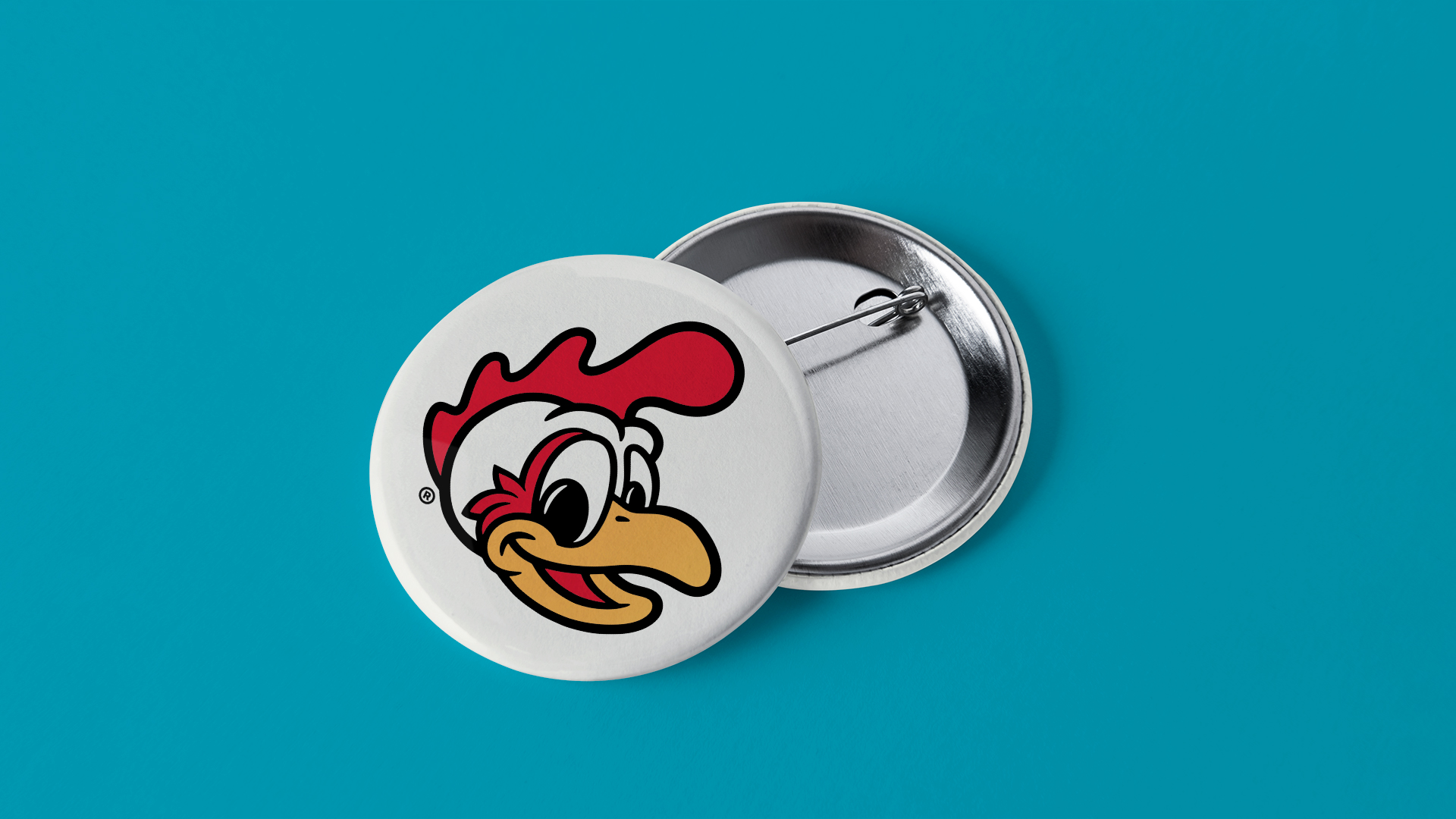 A round pin badge featuring a cartoon chicken head with a white face, red comb, and yellow beak. The badge is set against a turquoise background.