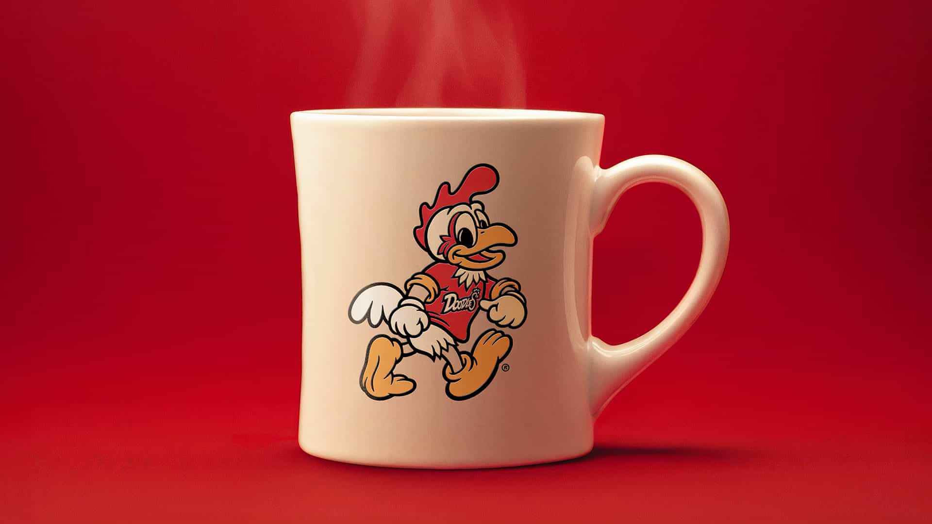 A white ceramic mug with steam rising from it, featuring a colorful cartoon chicken character in a dynamic pose, wearing a red shirt with "Doodles" written on it. The mug is set against a solid red background, emphasizing the playful design.