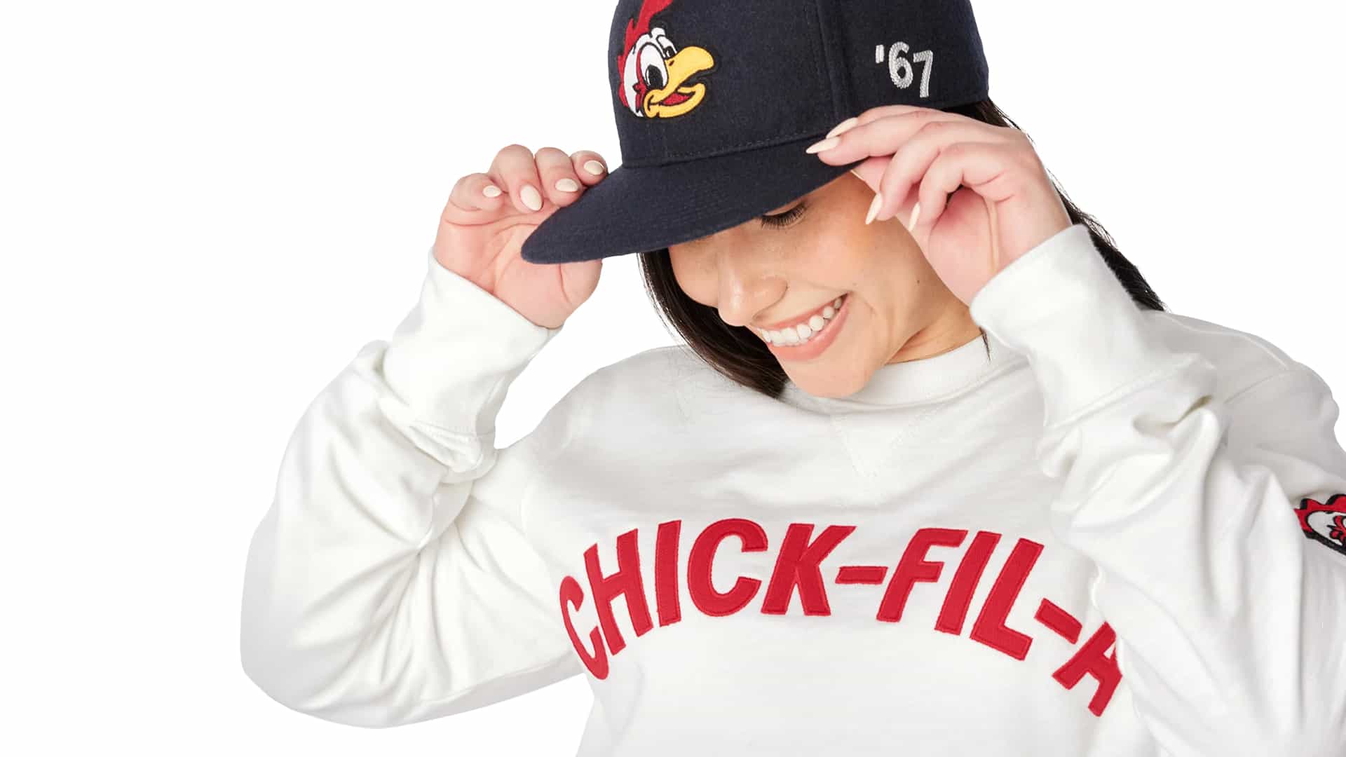A person wearing a white sweatshirt with "CHICK-FIL-A" written on it in red letters. They are also wearing a black baseball cap with a cartoon chicken logo on the front and the number "67" on the side. The person is adjusting the cap with both hands.